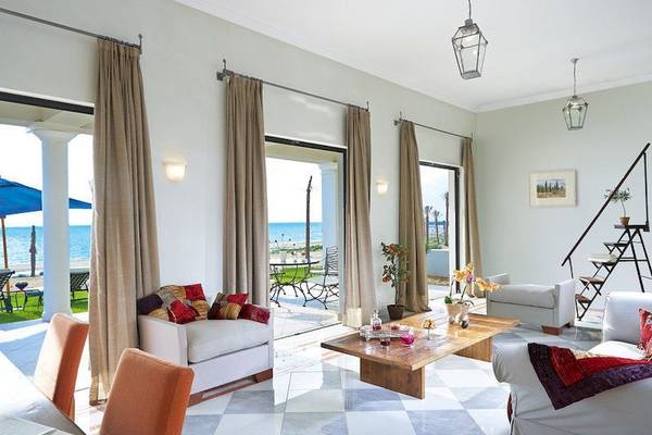 Wohnzimmer des Grecotel Olympia Oasis Aqua Park mit Blick auf das Meer und Möbeln vor großen Glastüren.