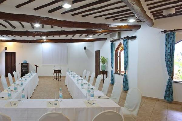 Ein Konferenzraum im Bluebay Beach Resort & Spa mit zwei langen Tischen, weißen Stühlen und blauen Vorhängen.