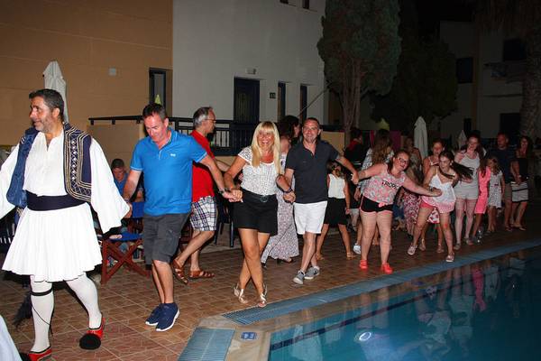 Gäste im Blue Aegean Hotel & Suites tanzen am Pool, begleitet von einem Mann in traditioneller griechischer Kleidung.