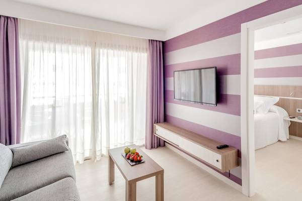 Innenansicht einer Hotelsuite im Barcelo Fuerteventura Mar mit lila und weißen Wänden, einem Sofa, einem Tisch und einem Fernseher.
