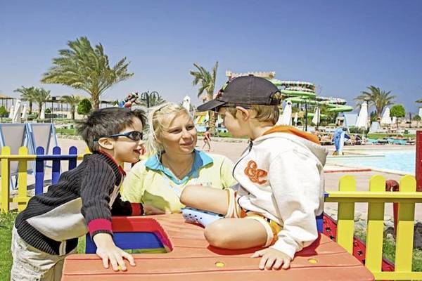 Zwei Kinder und eine Frau spielen auf einem Spielplatz im Freien, mit einem Schwimmbad im Hintergrund.
