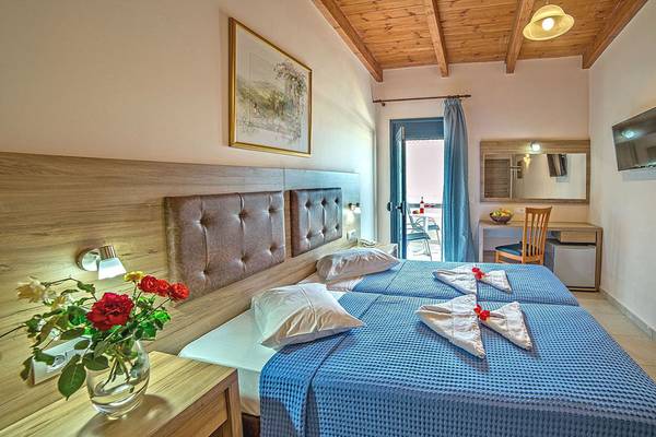 Zwei Betten im Zimmer im Blue Aegean Hotel & Suites, beide mit blauen Laken und weißen Kissen, ein Blumenstrauß in einem Glas und ein Fernseher an der Wand.