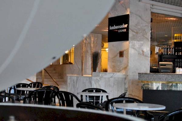 Innenansicht der Ambassador Lounge im AX Odycy Hotel mit weißen Tischen und Stühlen, Marmorwänden und einer Theke.
