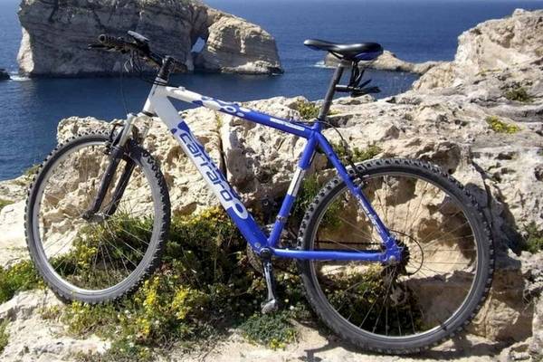 Ein blau-weißes Mountainbike steht auf einem Felsvorsprung am Meer, im Hintergrund eine beeindruckende Felsformation.