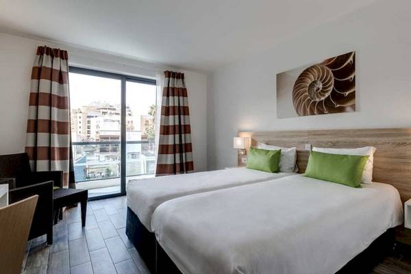 Doppelzimmer im AX Odycy Hotel mit zwei Betten, grünen Kissen, einem Balkon und Stadtblick.