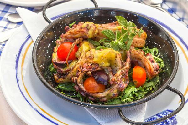 Gegrillte Oktopus-Speisen in einer schwarzen Schüssel auf einem Teller, umgeben von Salat und Tomaten.
