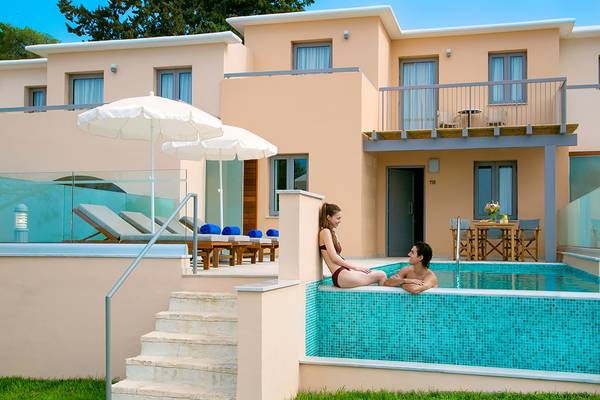 Ein Paar sitzt im Pool einer Villa, neben der es Sonnenliegen und ein großes Gebäude gibt.