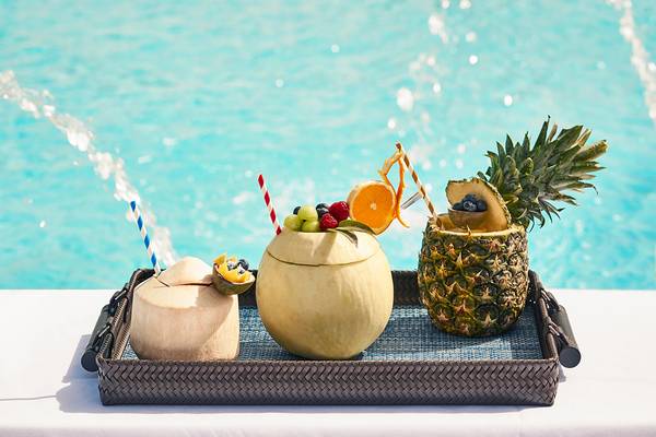 Cocktails in geschnittenen Kokosnuss- und Ananasschalen auf einem Tablett neben einem Pool im Raffles The Palm Dubai