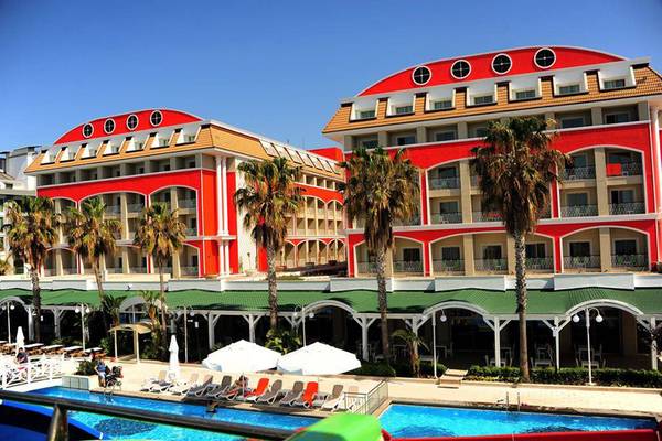 Außenansicht des Orange County Belek - Family Concept Hotels mit roten Gebäuden, grünen Dächern und Palmen vor dem Pool.
