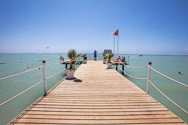 Ein Holzsteg am Strand des Orange County Belek Family Concept Hotels mit einem Mann, der auf die Kamera blickt.