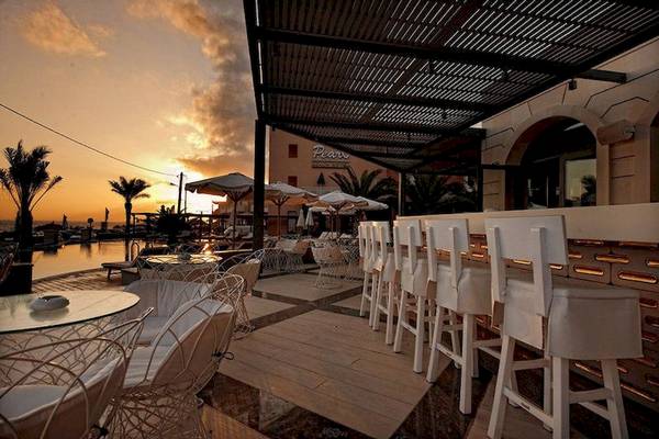 Außenbereich des Aegean Pearl Hotels mit Terrassenstühlen, Tischen und Sonnenuntergang im Hintergrund.