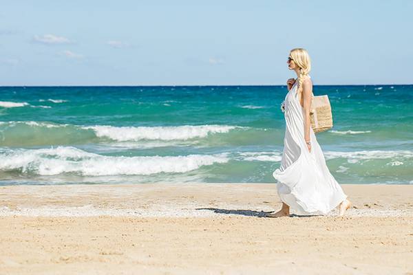 Eine Frau in einem weissen Kleid geht mit einer Tasche am Strand entlang.