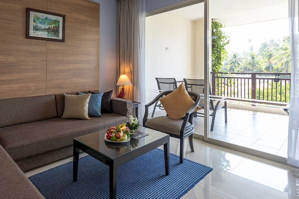 Wohnzimmer des Kantary Beach Hotel mit Sofa, Tisch, Balkon und Ausblick auf eine Gartenlandschaft.