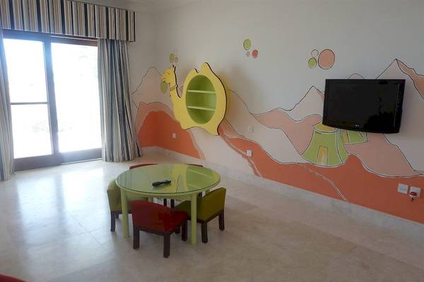 Juweira Boutique Hotel Kinderzimmer mit grünem Tisch, roten und grünen Stühlen, Fernseher und farbenfroher Wandgestaltung.