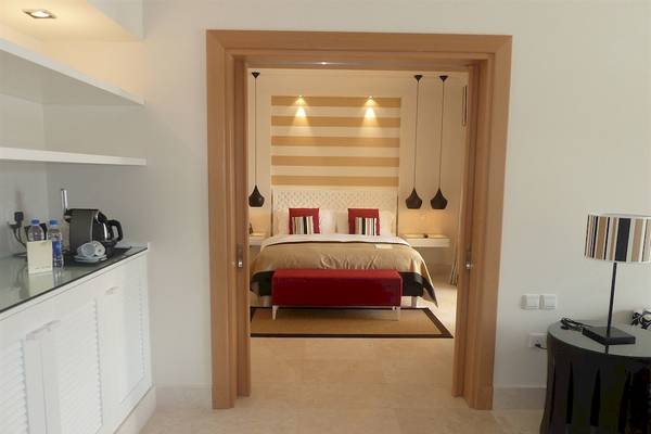 Innenansicht des Schlafzimmers im Juweira Boutique Hotel mit Doppelbett, rotem Hocker und modernen Lampen.