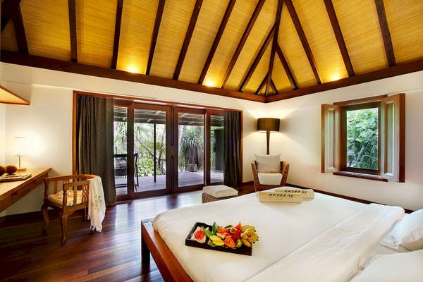Innenansicht eines Schlafzimmers im Gangehi Island Resort mit Früchten auf dem Bett und Panoramafenstern.