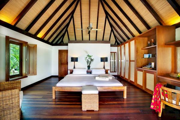Innenansicht eines Schlafzimmers im Gangehi Island Resort mit Holzboden, Bett, Nachttischen und Deckenventilator.