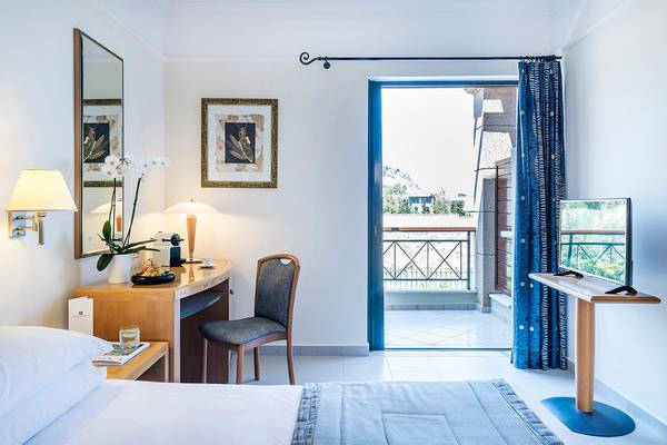 Innenansicht eines Zimmers im Mitsis Lindos Memories Resort & Spa mit Bett, Schreibtisch, Stuhl, Balkonzugang und Fernseher.