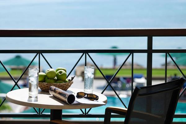 Ein Balkon mit einem Tisch, auf dem eine Zeitschrift, eine Sonnenbrille, ein Glas Wasser und ein Korb mit Äpfeln steht. Im Hintergrund ist ein Pool zu sehen.