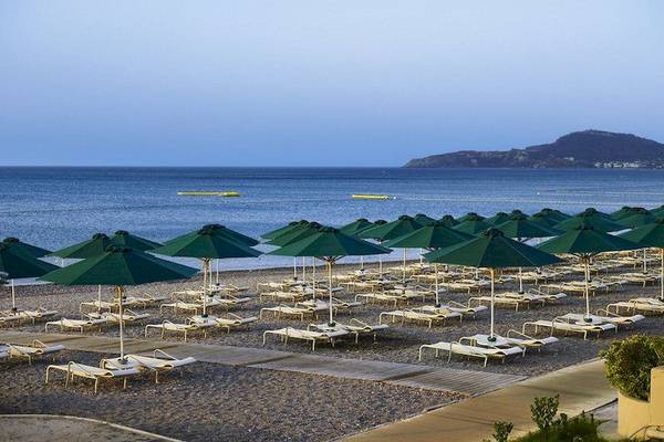 Foto der Strandseite des Esperos Village Blue & Spa Hotels mit vielen Sonnenliegen und grünen Sonnenschirmen.