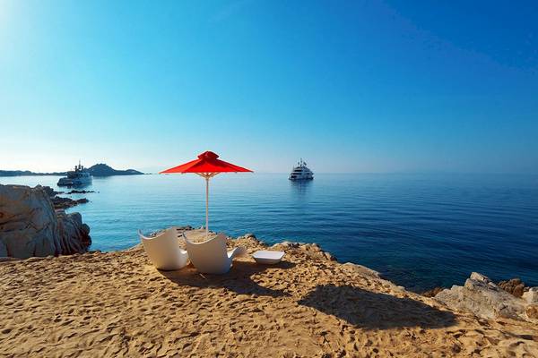 Petasos Strand mit roter Sonne, weißen Sesseln und zwei Booten im Hintergrund.