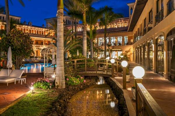 Abendansicht des Secrets Bahia Real Resort & Spa mit beleuchtetem Gehweg, Brücke und Swimmingpool.
