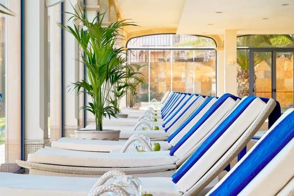 Reihe von weißen und blauen Sonnenliegen im Innenbereich des Secrets Bahia Real Resort & Spa mit Pflanzen und Glasfenstern im Hintergrund.