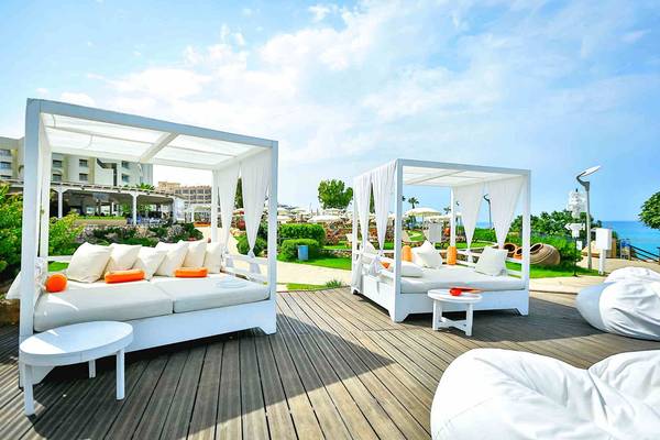 Zwei weiße Loungebetten mit Vorhängen auf einer Holzterrasse am Capo Bay Hotel mit Blick auf das Meer.