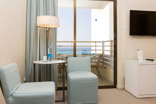 Hotelzimmer mit Meerblick und Balkon, ausgestattet mit einem Tisch, Stühlen, einer Lampe und einem Fernseher.
