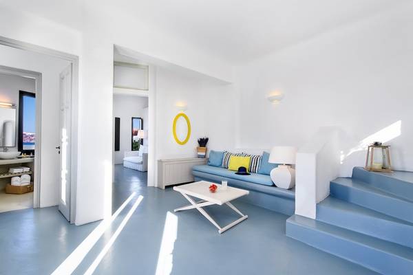 Innenansicht des Ambassador Aegean Luxury Hotel & Suites mit einer offenen Tür, einem Wohnzimmer mit einem weißen Tisch, einem blauen Sofa, einer weißen Lampe und einer gelben Lampe.