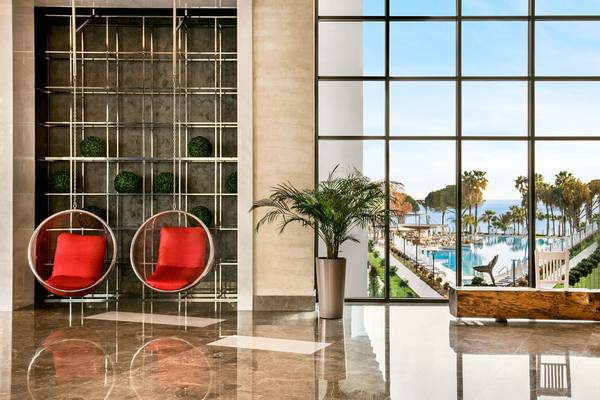 Eingangsbereich eines Hotels mit zwei roten Sessel und einer großen Fensterfront, die einen Blick auf den Pool und das Meer bietet.