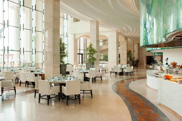 Innenbereich des St. Regis Saadiyat Island Resorts mit weißen Tischen und Stühlen, Glaswänden und Pflanzen.
