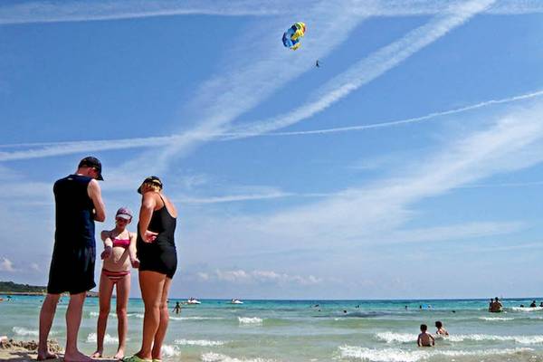 Strandszene mit mehreren Personen, die auf das Meer schauen, und einem Segelflugzeug am Himmel.