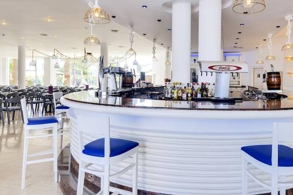 Die Bar im Tropic Garden Hotel Apartments mit weißen Stühlen, blauen Sitzen und einer Theke, auf der Getränkeflaschen und ein Coca-Cola-Kühlschrank stehen.