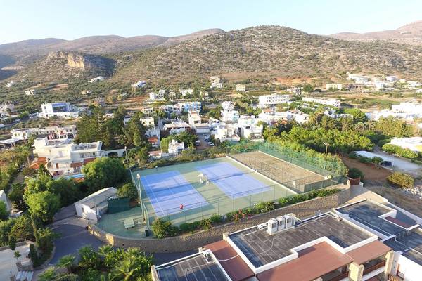 Luftaufnahme des Tennisplatzes im Ikaros Beach Luxury Resort & Spa mit Gebäuden und Bergen im Hintergrund.