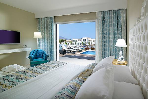 Schlafzimmer mit Bett, offenem Fenster und Aussicht auf einen Pool im Anemos Luxury Grand Resort.