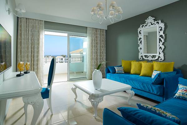 Innenansicht des Wohnzimmers im Anemos Luxury Grand Resort mit blauem Sofa, weißem Tisch und Aussicht auf den Balkon.