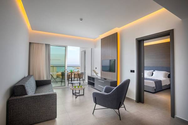 Innenansicht eines Zimmers im Cavo Maris Beach Hotel mit Sofa, Tisch und Fernseher.
