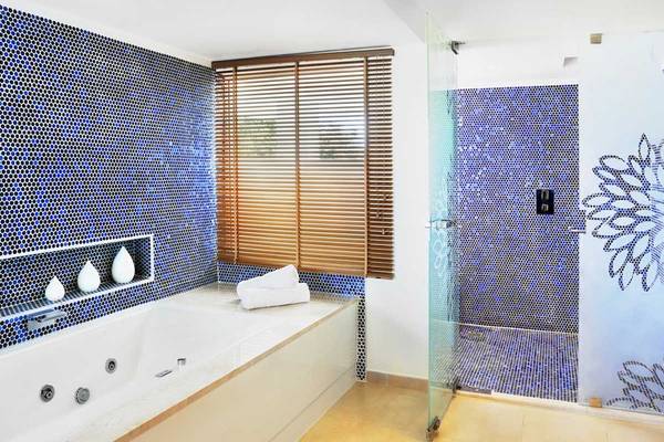 Ein Badezimmer mit blauen Fliesen, Badewanne, Dusche und Fenster mit Jalousien.