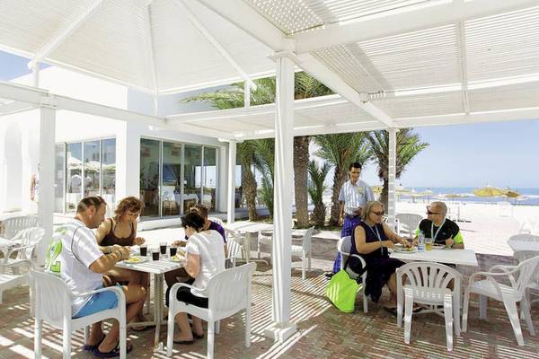 Personen sitzen an Tischen in einem Restaurant unter einem Dach mit Blick auf den Strand.