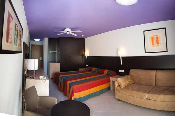 Innenansicht eines Hotelzimmers im SBH Monica Beach Resort mit Bett, Sofa, Schreibtisch und Dekor.
