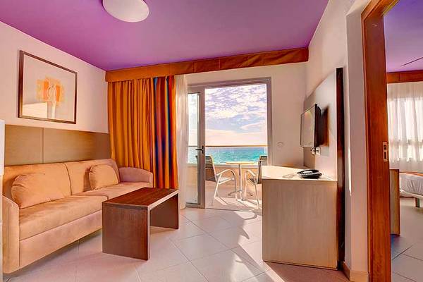 Innenansicht eines Zimmers im SBH Monica Beach Resort Hotel mit Sofa, Tisch und Balkon.