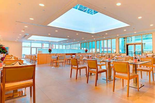 Innenansicht des Restaurants im SBH Monica Beach Resort mit runden Tischen und Stühlen, hoher Decke und Glasfenstern.