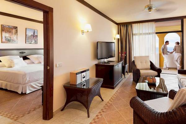 Innenansicht einer Suite im Occidental Jandia Royal Level Hotel mit Schlafzimmer, Wohnzimmer und Balkon.