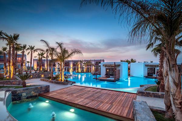 Abendliche Ansicht des Swimmingpools im Stella Island Luxury Resort & Spa mit Holzbrücke und beleuchteten Palmen.