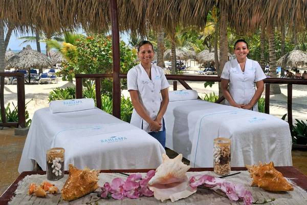 Zwei Mitarbeiter im Bahia Spa des Grand Bavaro Hotels stehen neben zwei Massagebetten, die mit weißen Handtüchern und Muscheldekor auf einem Tisch vorbereitet sind.