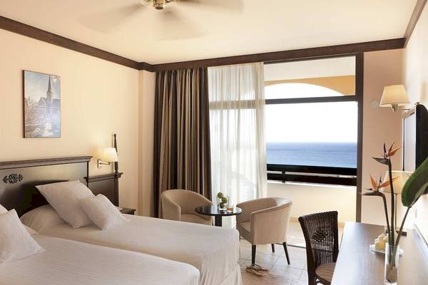 Doppelzimmer mit Meerblick im Occidental Jandia Playa Hotel, mit zwei Betten, Sesseln, einem Tisch und einem großen Fenster mit Balkon.
