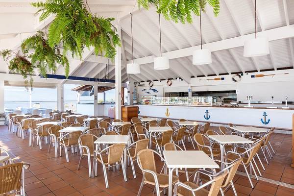 Innenansicht des Restaurants des Landmar Costa Los Gigantes Hotels mit vielen Tischen und Stühlen, hängenden Pflanzen und einer Theke im Hintergrund.