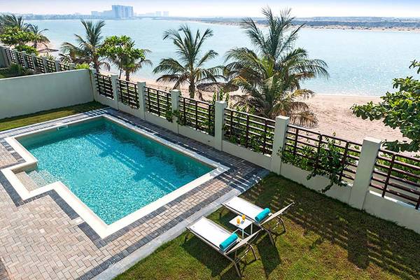 Ein Swimmingpool mit zwei Liegen und einem Tisch mit Getränken auf einer Terrasse mit Blick auf den Strand und das Meer, im Hintergrund Hotelgebäude.