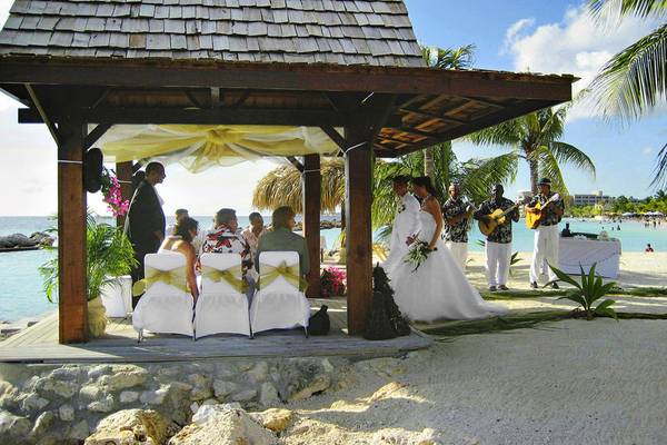 Hochzeitszeremonie am Strand vor einem Pavillon mit Musikern, die Gitarre spielen.